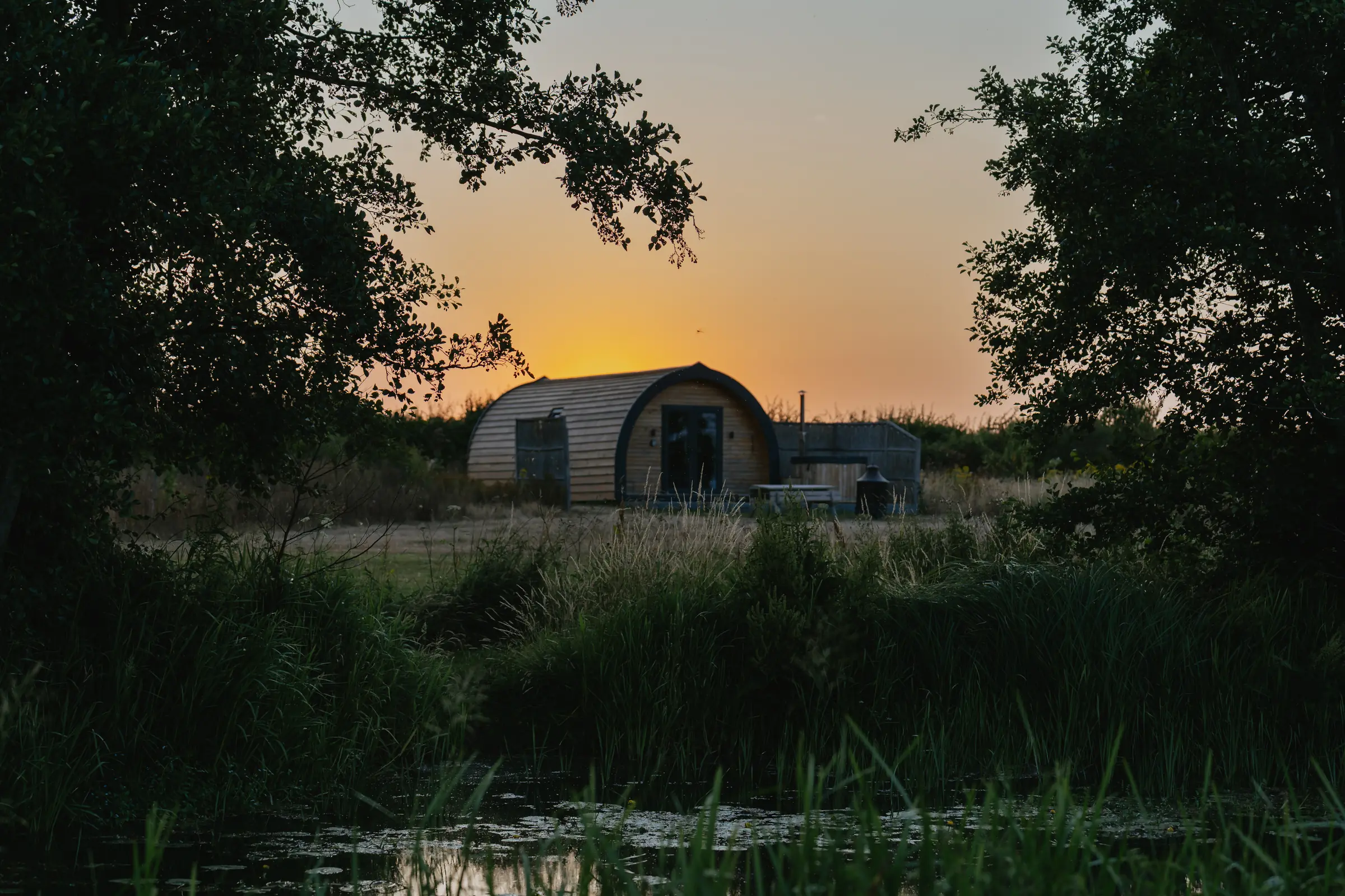 A timber pod beside the water at sunset, framed by trees